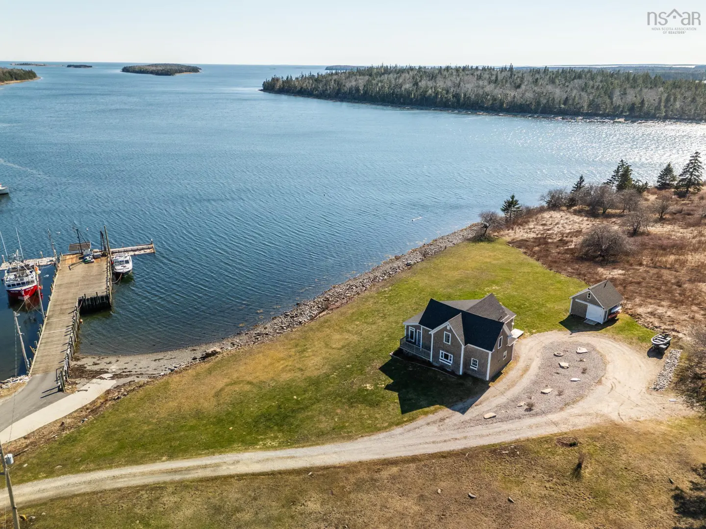 Aerial view of a gray house with a dock on the water. Boats are docked at the pier. A detached garage sits near the house.