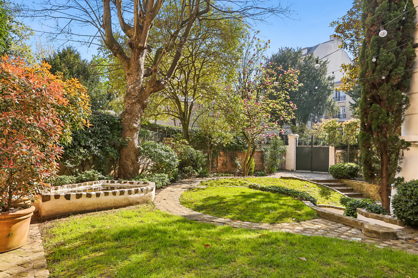 A lush backyard garden with a stone-bordered lawn, a small pond, mature trees, and string lights under a clear blue sky.
