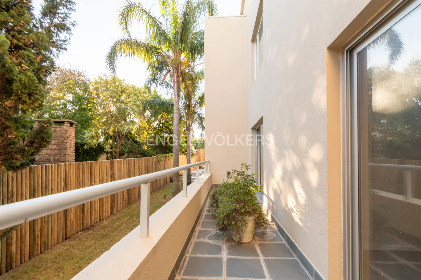 Balcony view with gray tile floor, white railing, and potted plant. A wooden fence and green trees are in the background.