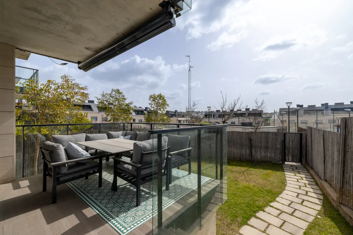 Modern terrace with gray outdoor furniture, patterned rug, and glass railing overlooking a small garden with stone path.