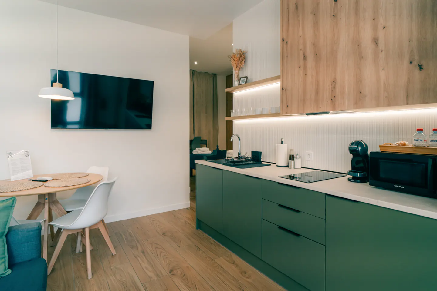 Interior view of a modern apartment with green cabinets, wood floors, a round table, and a TV.