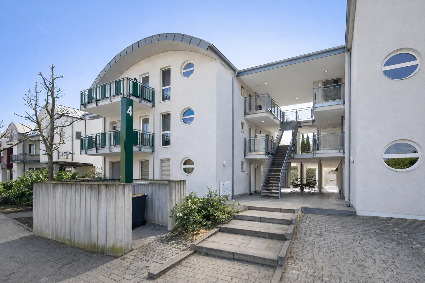Exterior of a modern, white apartment building with balconies and round windows under a blue sky. A staircase leads to upper floors.