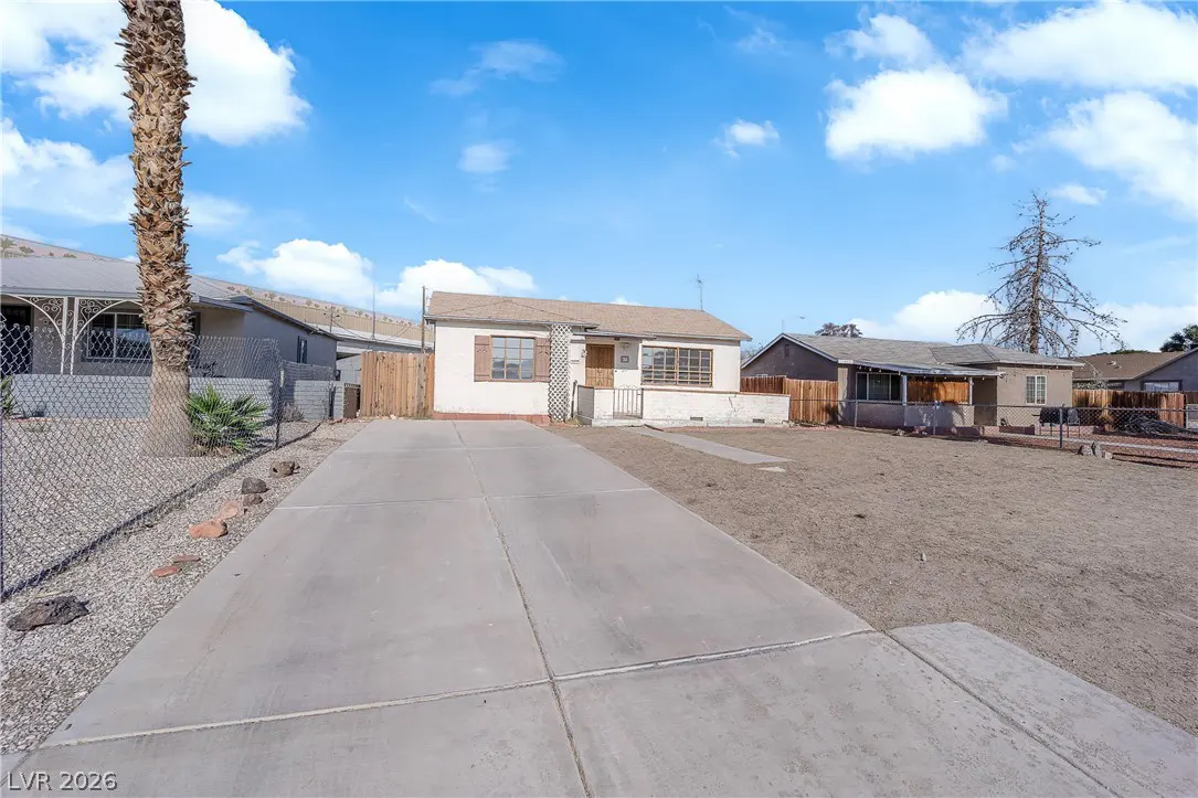 A single-story beige house with a concrete driveway under a blue, cloudy sky. Neighboring houses and a tall palm tree are visible.