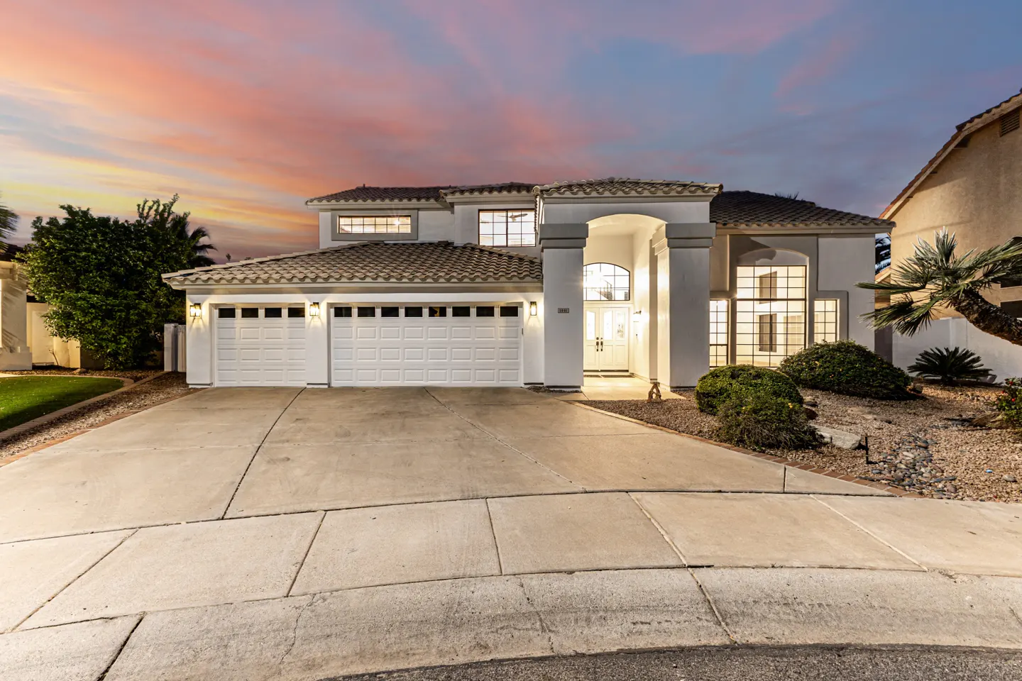 Two-story white house with a three-car garage and a tile roof at dusk. A concrete driveway leads to the house.