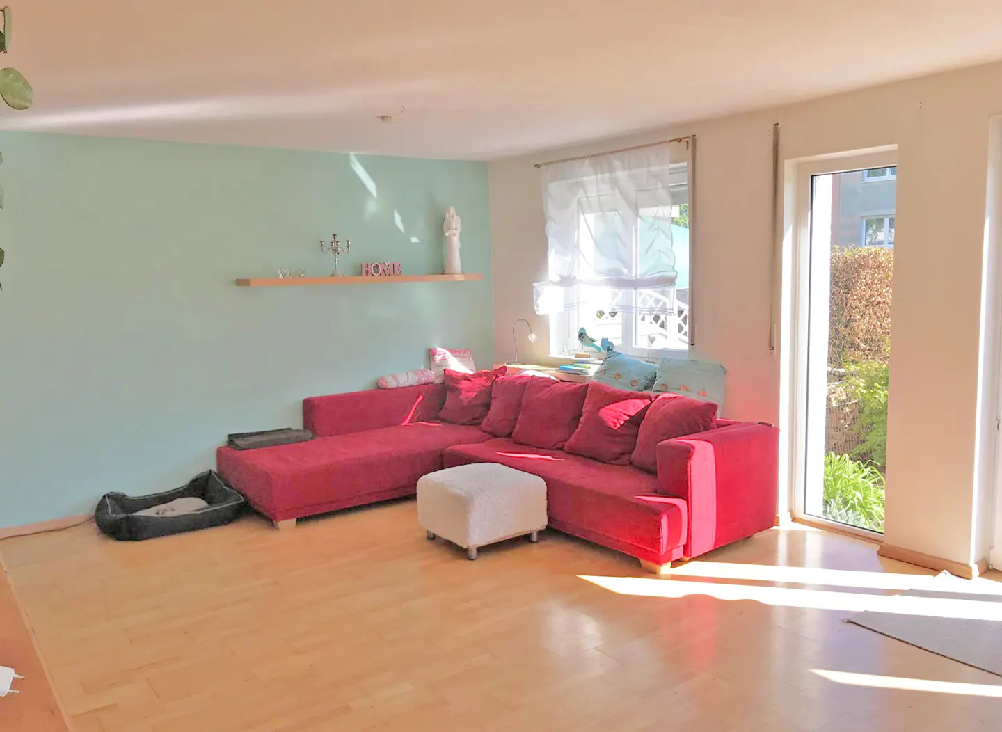 Bright living room with a red sectional sofa, white ottoman, and light wood floors. A dog bed sits near a light green wall with a shelf. Sunlight streams through a window.