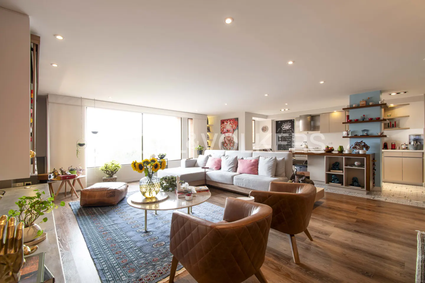Open-concept living room with a gray sofa, brown leather chairs, and a blue patterned rug. A kitchen is visible in the background.