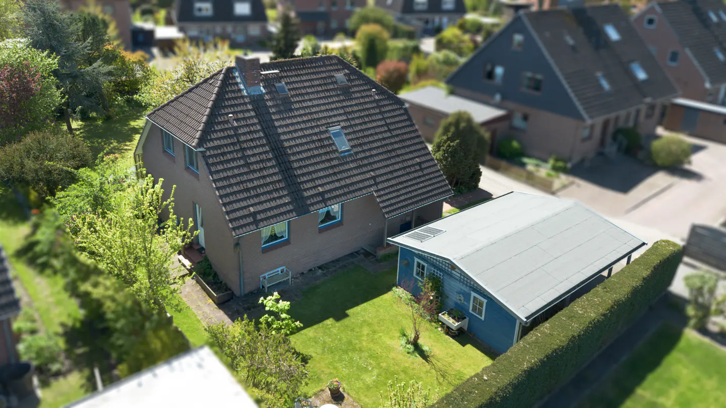 Aerial view of a brown house with a dark tiled roof, a blue shed, and a green backyard in a residential neighborhood.