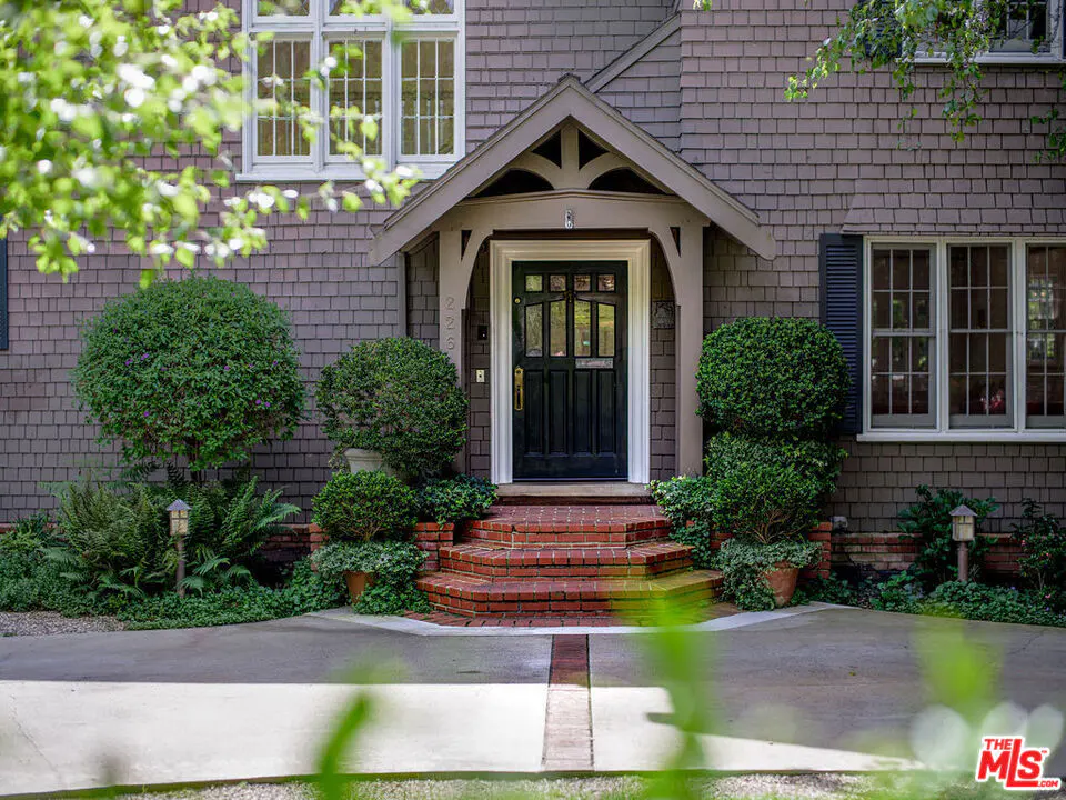 Front view of a brown shingle house with a black front door and brick steps. Green bushes flank the entrance.
