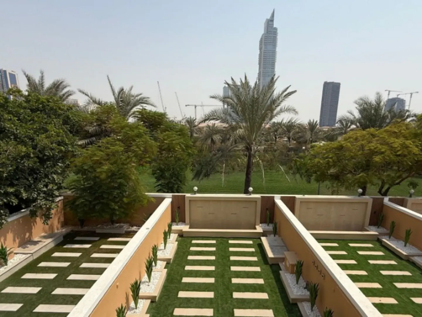A manicured backyard with grass, stone pavers, and beige walls, with palm trees and city buildings in the background.