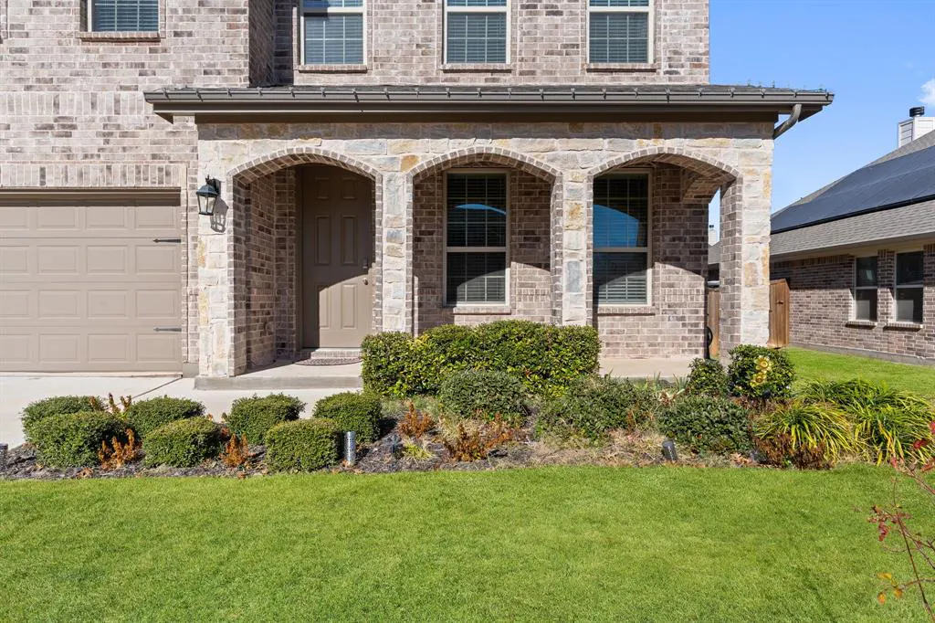 Brick house with arched porch, green lawn, and trimmed bushes. A tan garage door is on the left. Windows are visible above the porch.