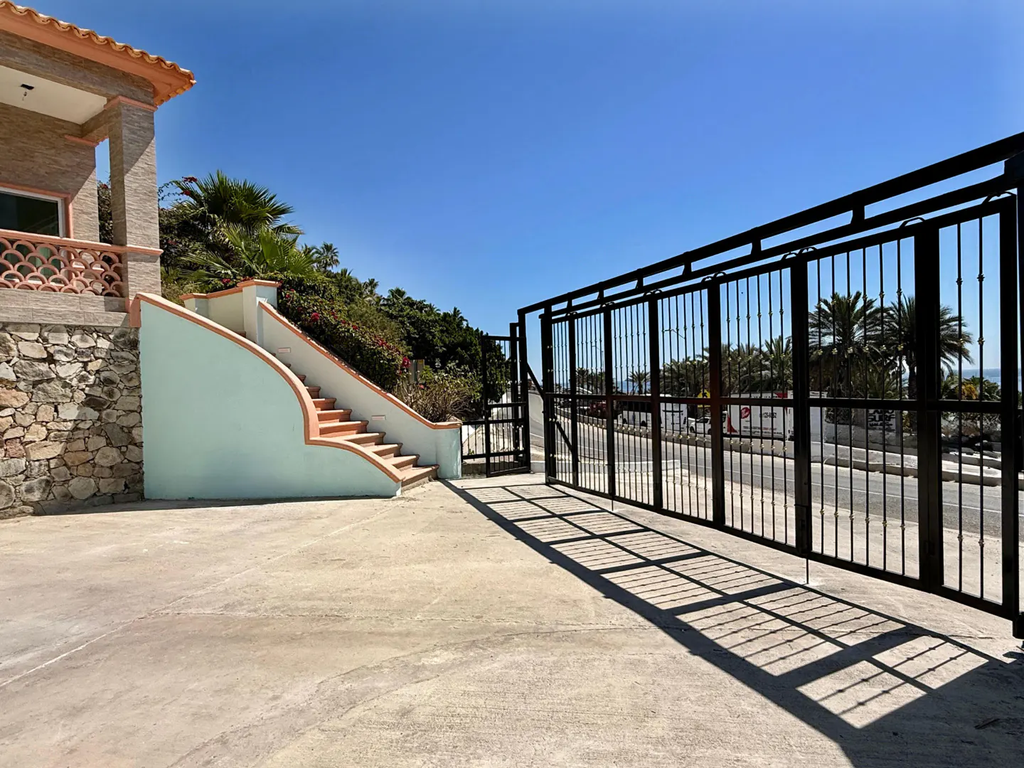 Exterior view of a property with a black metal gate, stairs with light blue walls and peach trim, and lush greenery under a clear blue sky.