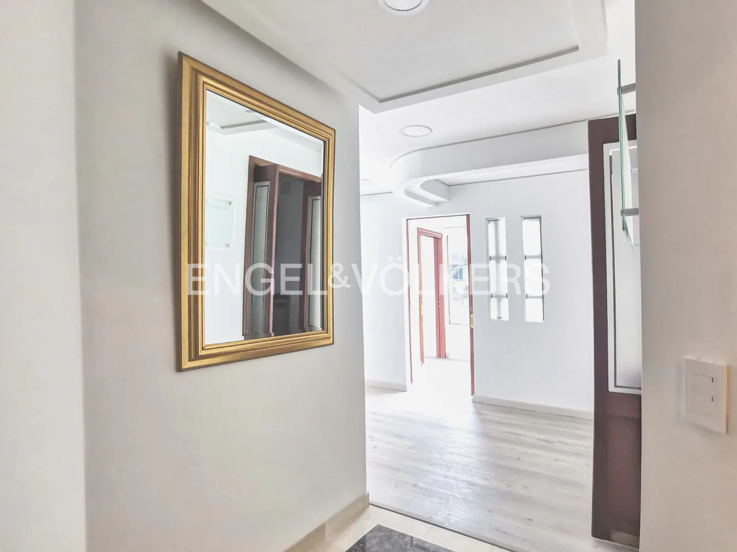 Entryway with a gold-framed mirror on a white wall, reflecting a doorway. A hallway with white walls and light wood floors leads to other rooms.