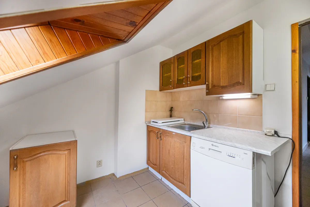 A small kitchen with wood cabinets, white countertops, and a white dishwasher. The ceiling is angled with wood paneling.