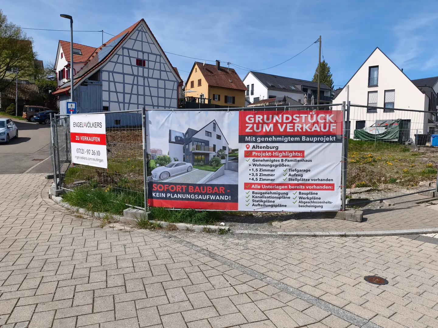 A fenced lot with "Land for Sale" signs in German, featuring a modern house rendering, in a residential area.