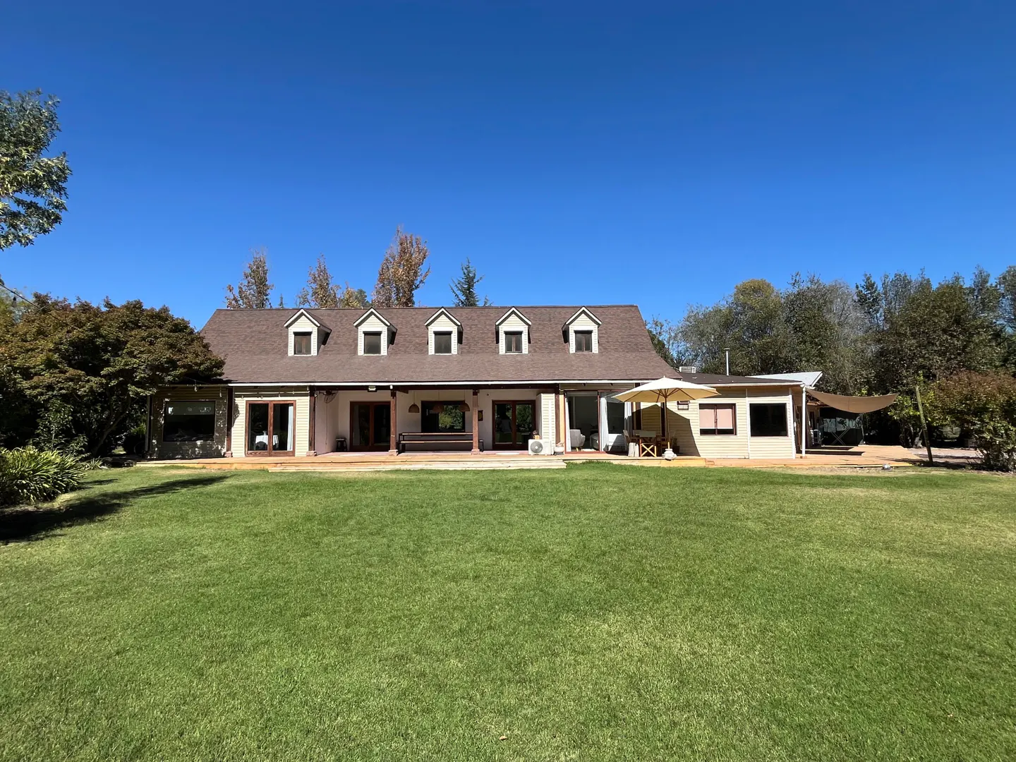 Wide shot of a tan house with a brown roof, dormer windows, and a large green lawn under a clear blue sky.