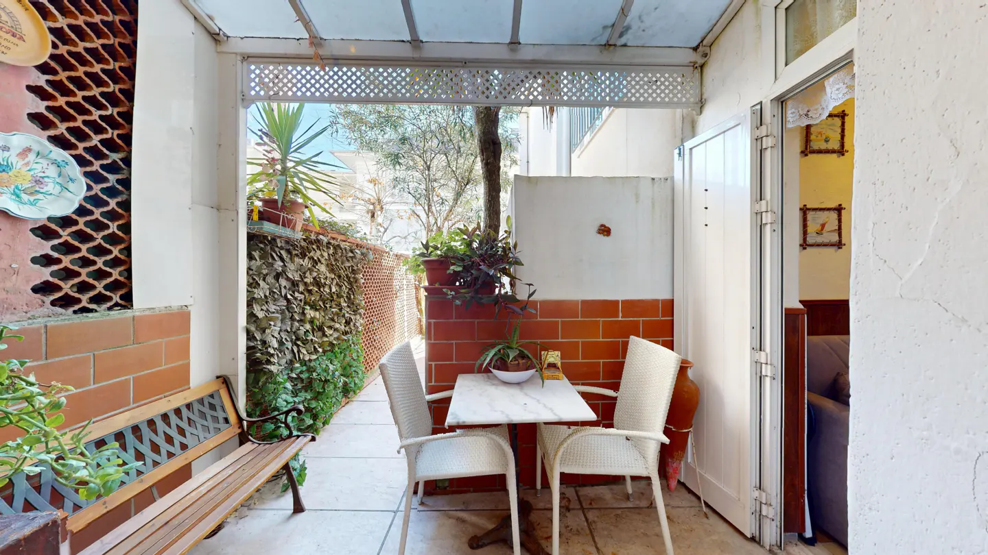 Outdoor patio with a marble table and two white chairs. A wooden bench sits against a brick wall with greenery.