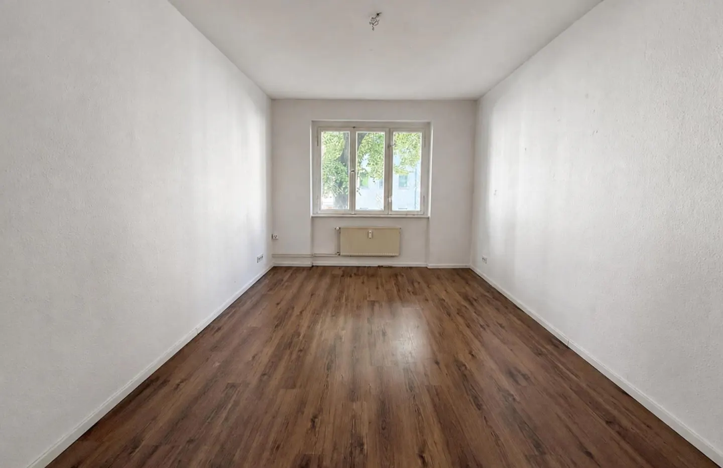 Empty room with white walls, wood-look laminate flooring, a central window, and a radiator underneath.