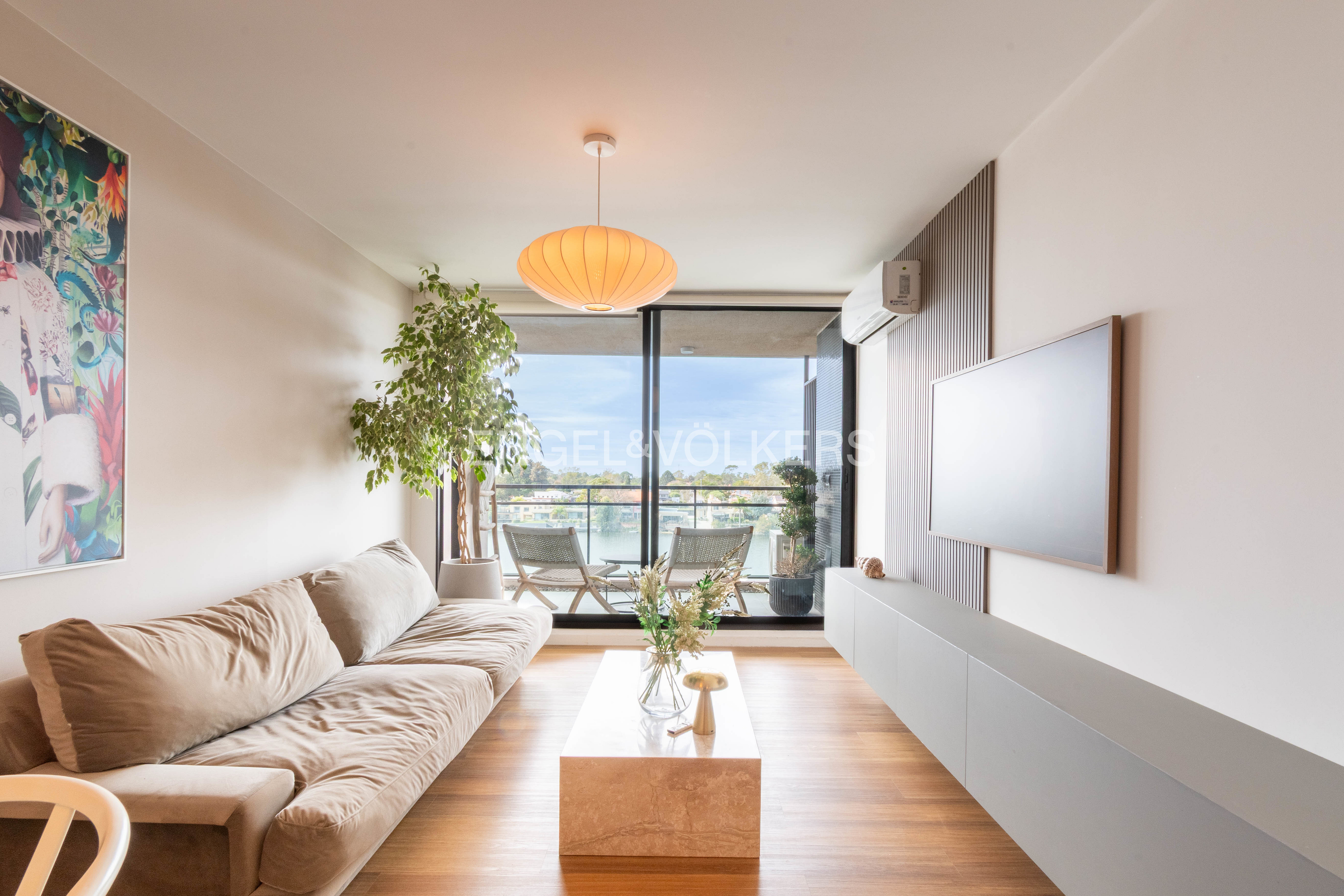 Living room with a beige sofa, marble coffee table, and a balcony overlooking a lake. A large painting hangs on the wall.