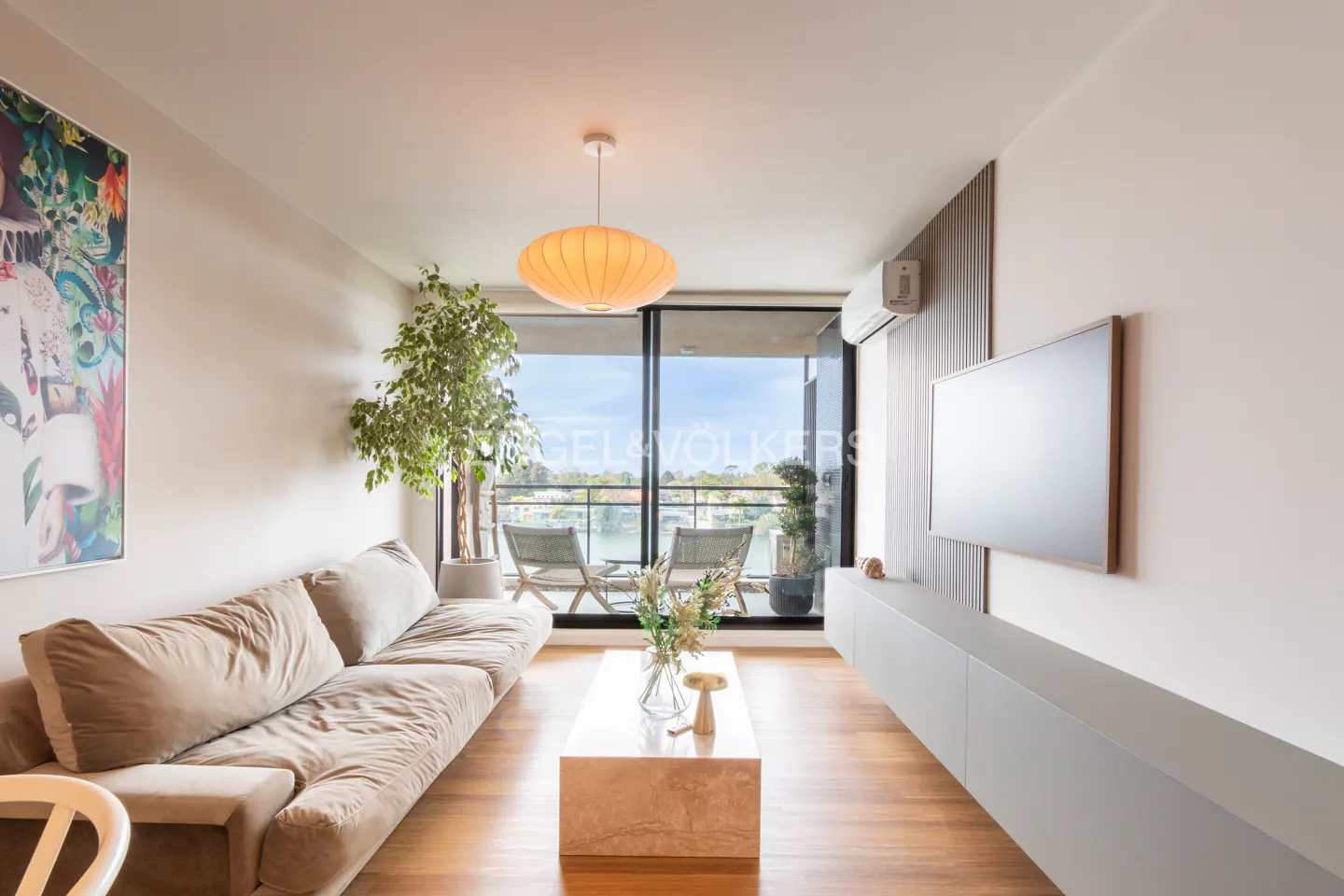 Living room with a beige sofa, marble coffee table, and a balcony overlooking a lake. A large painting hangs on the wall.
