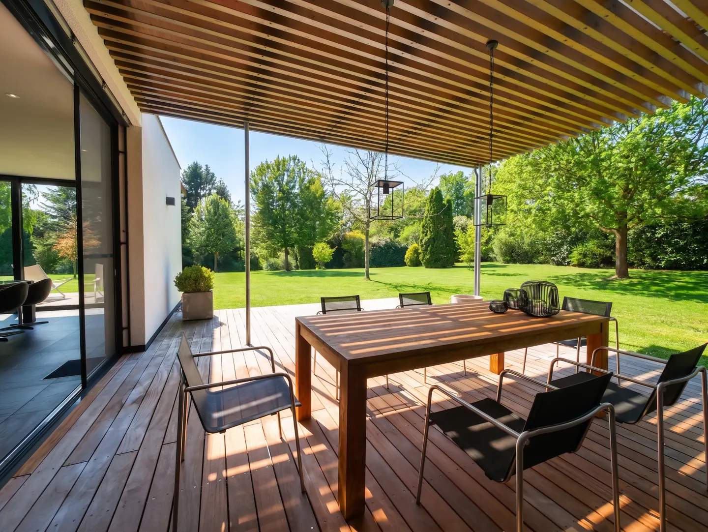 Outdoor dining area with a wooden table and chairs on a deck, under a slatted pergola, overlooking a green lawn.
