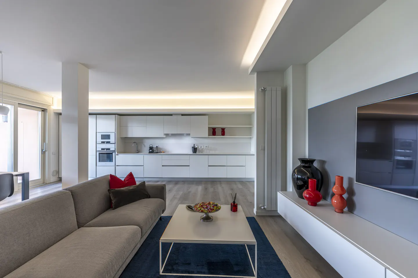 A modern open-concept living space with a gray sofa, white kitchen, and blue rug. A TV and vases are on a gray wall.