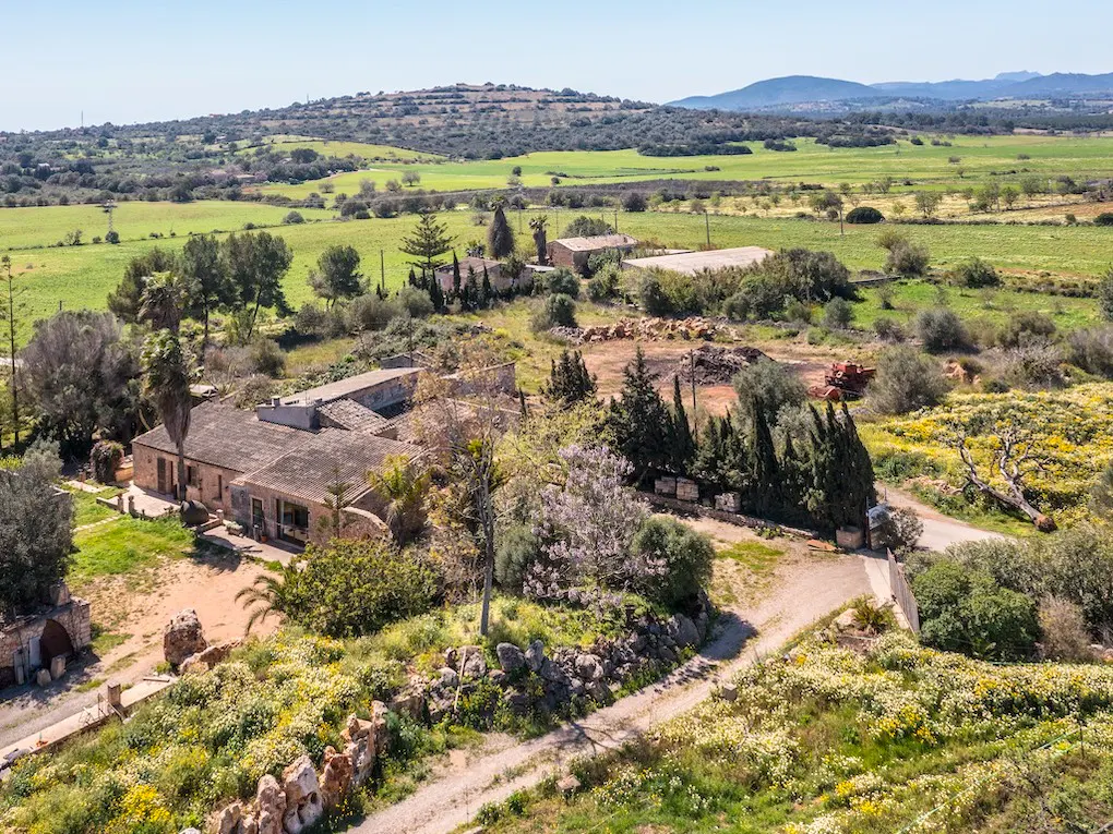 Aerial view of a stone farmhouse with a tiled roof, surrounded by green fields, trees, and a dirt road.