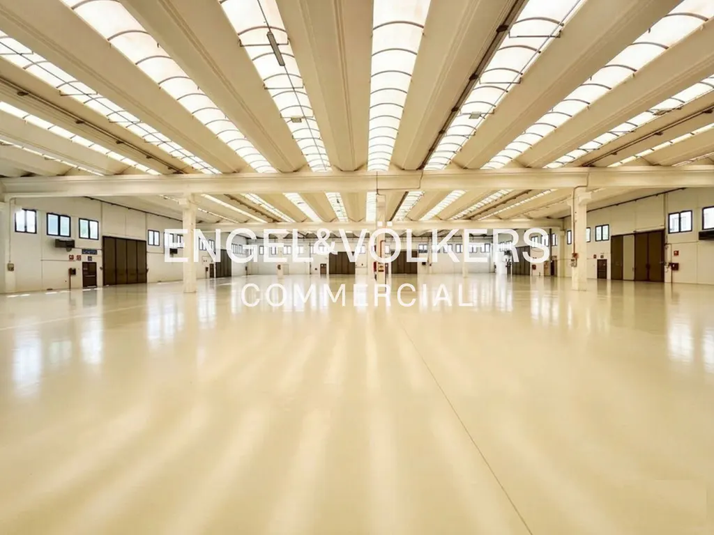 Wide, empty warehouse with a shiny beige floor and white ceiling with skylights. Doors and windows line the walls. "Engel & Volkers Commercial" overlay.