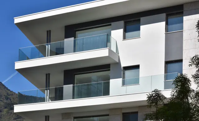 Modern white building with glass balconies and dark window frames, set against a blue sky and green trees.