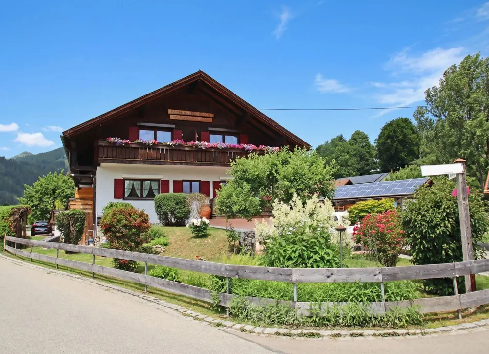 A two-story house with a brown roof, white walls, red shutters, and a balcony with flowers. A wooden fence surrounds the yard.