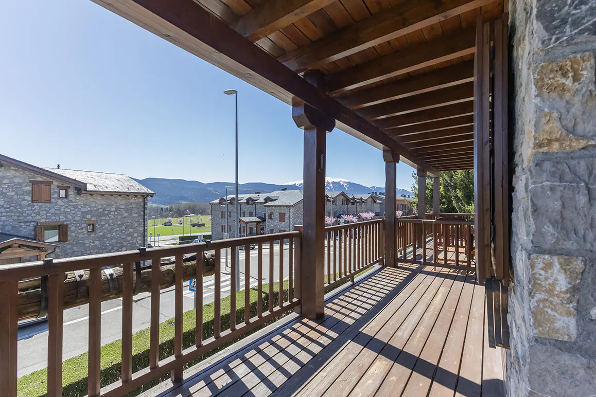 View from a wooden balcony with a brown railing, overlooking a street, buildings, and mountains in the distance.