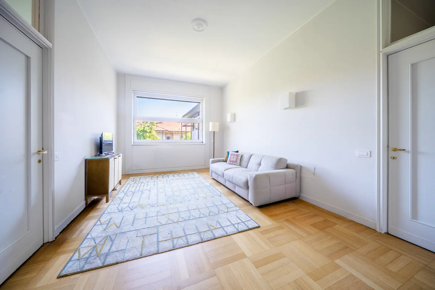 Bright living room with white walls, wood floors, and a geometric rug. A light gray sofa sits near a window with a view.