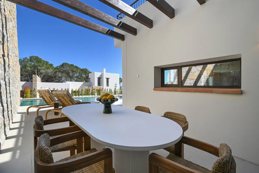 Outdoor dining area with white table, wicker chairs, and fruit bowl. Pool and lounge chairs visible in the background. Blue sky.