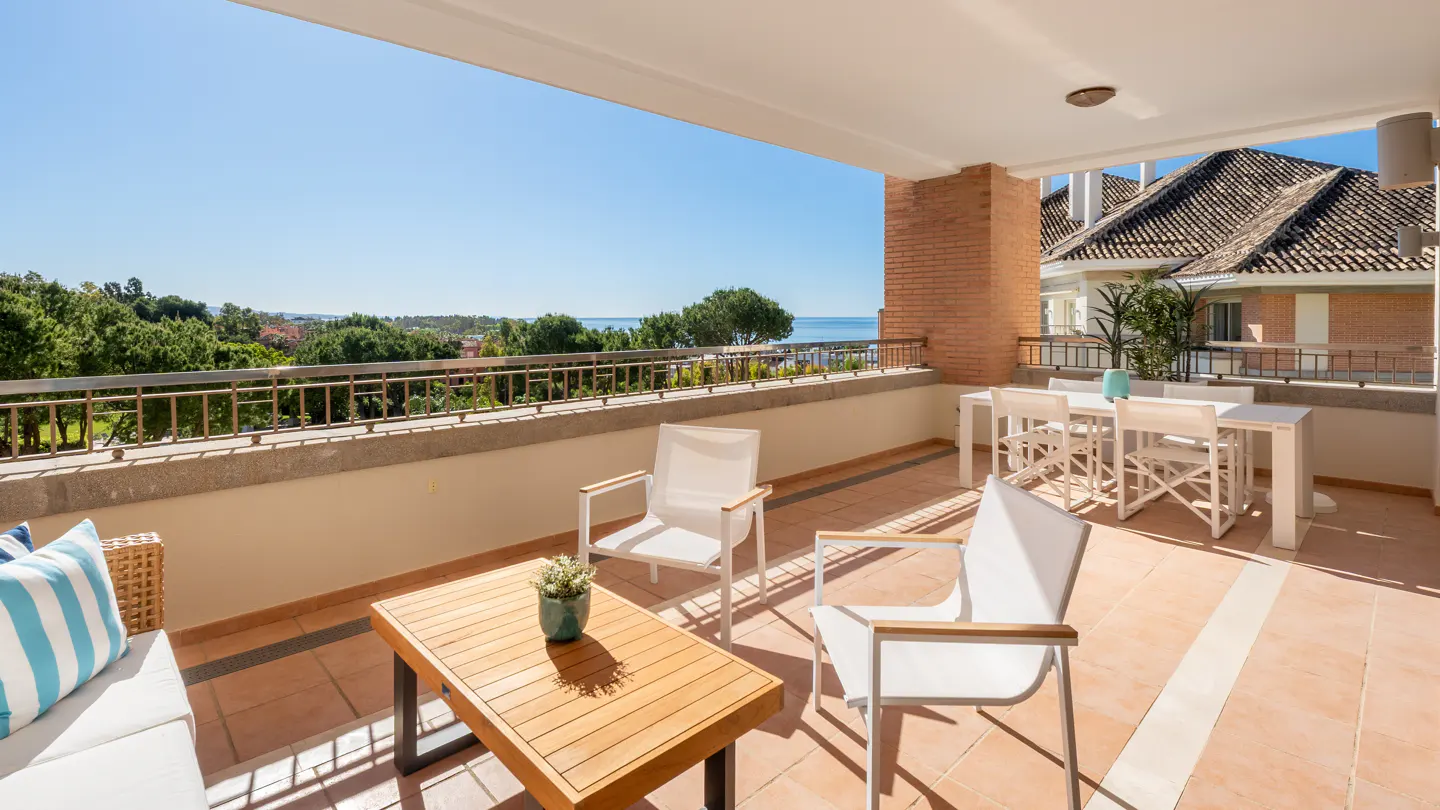 A sunlit balcony with terracotta tiles features white chairs, a wooden table, and a sofa with blue striped pillows, overlooking a scenic view.
