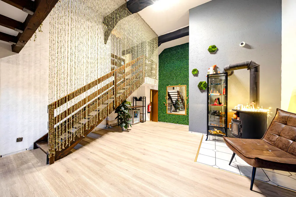 Interior view of a room with a wooden staircase, beaded curtains, and a green wall. A black stove and brown leather chair are visible.