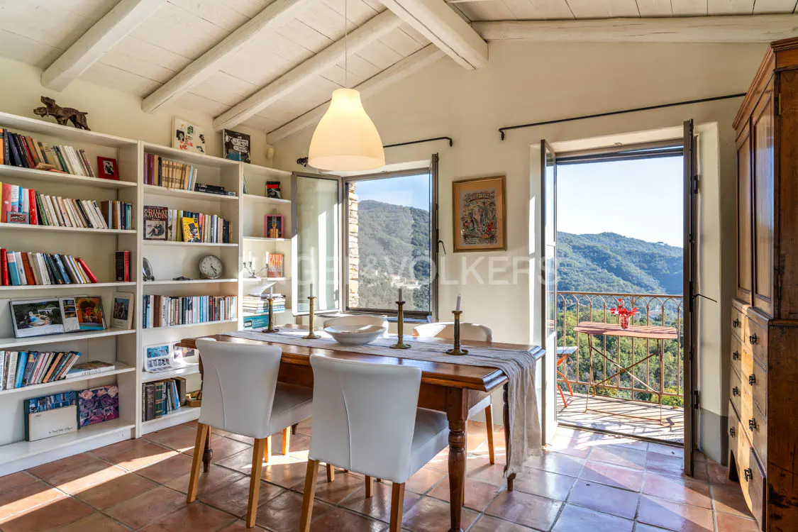 Bright dining room with terracotta tile floor, white bookshelves, and a balcony overlooking green hills.