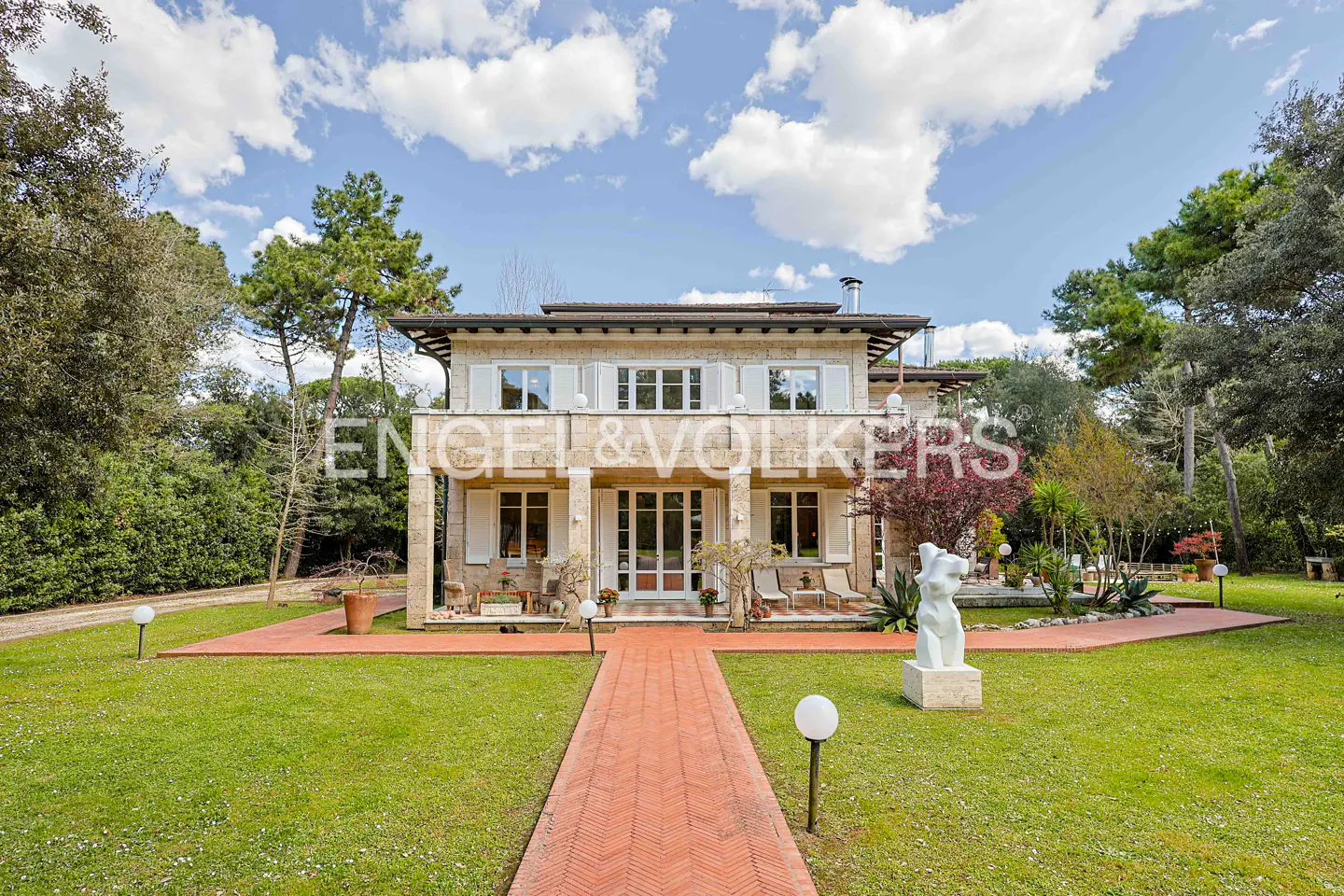 Two-story stone house with white shutters, a red brick walkway, and a green lawn under a blue sky with clouds. "Engel & Volkers" logo visible.