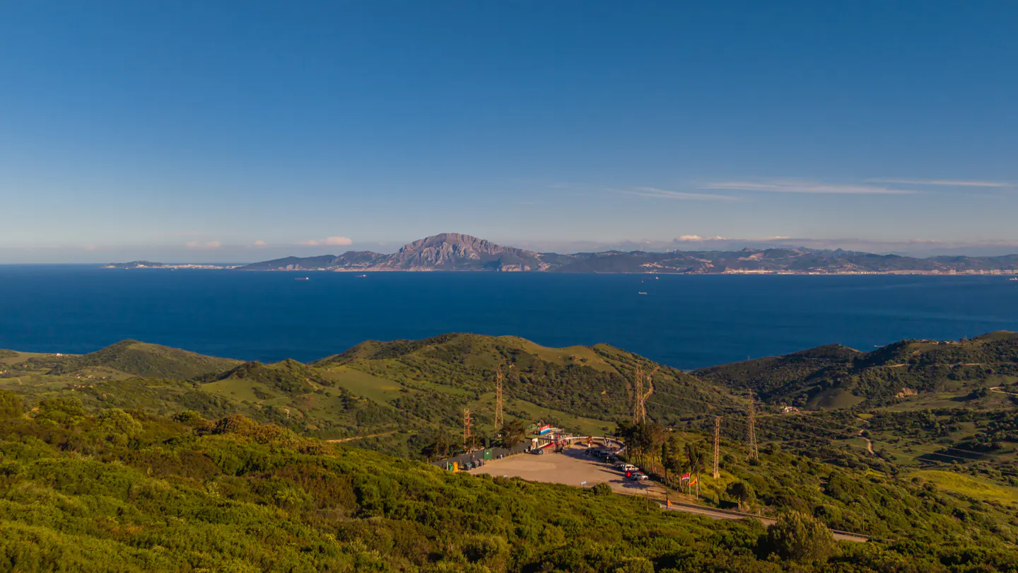 Scenic view of the Rock of Gibraltar across the sea, seen from green hills with a parking area and transmission towers.