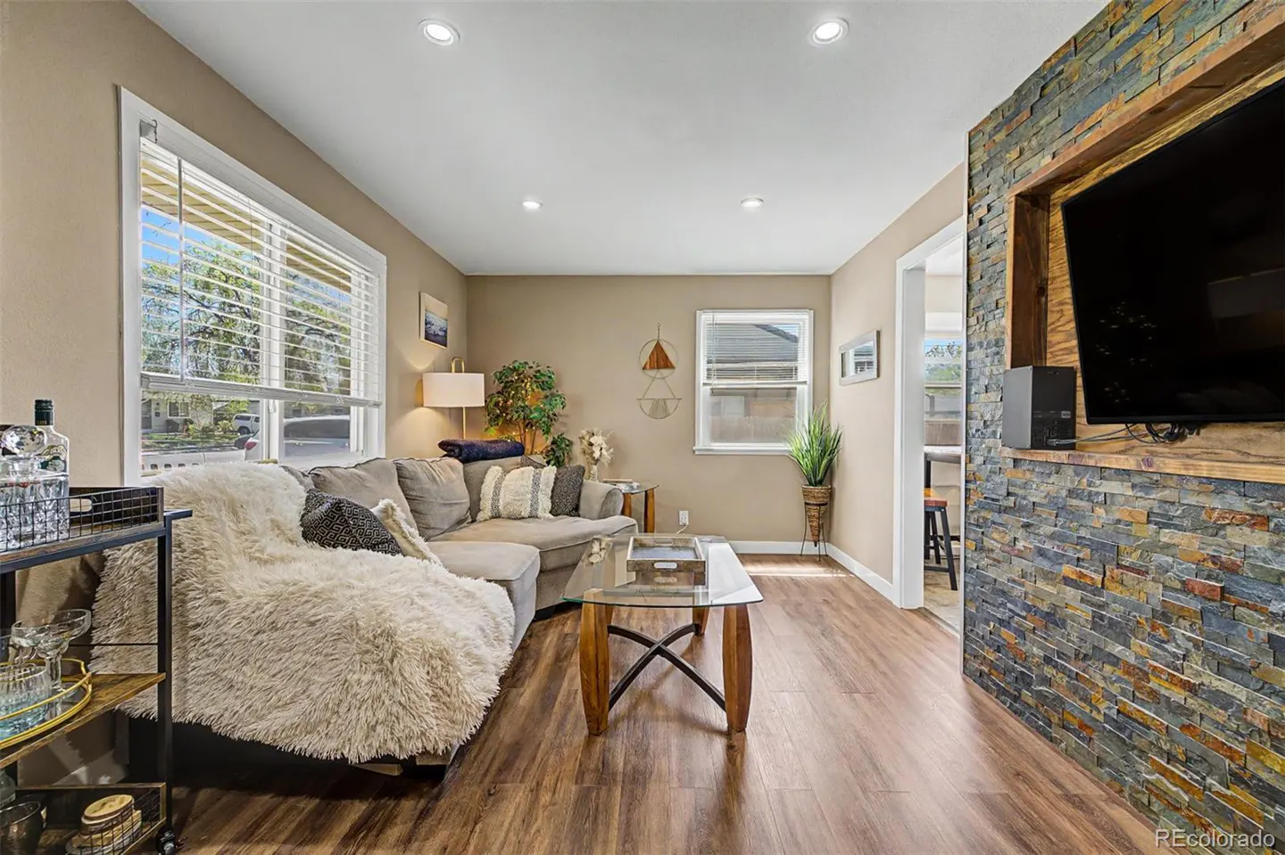 Living room with a gray sectional sofa, glass coffee table, and stone accent wall with a TV. Wood floors and neutral wall color.
