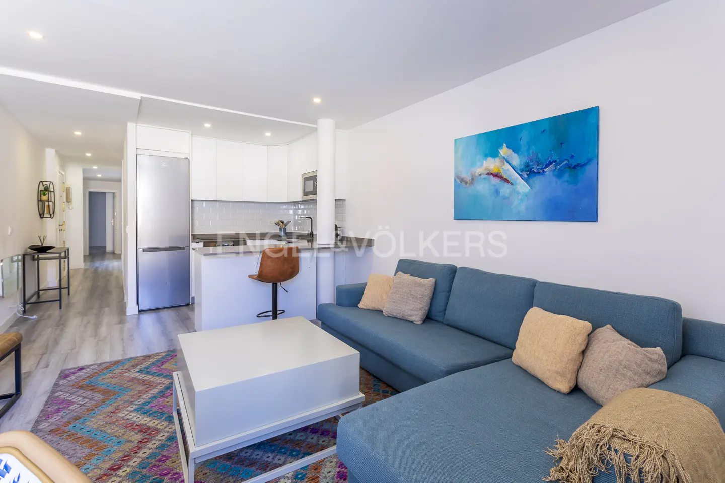 Bright, modern apartment interior with a blue sectional sofa, white kitchen, and colorful rug. Abstract art hangs on the white wall.