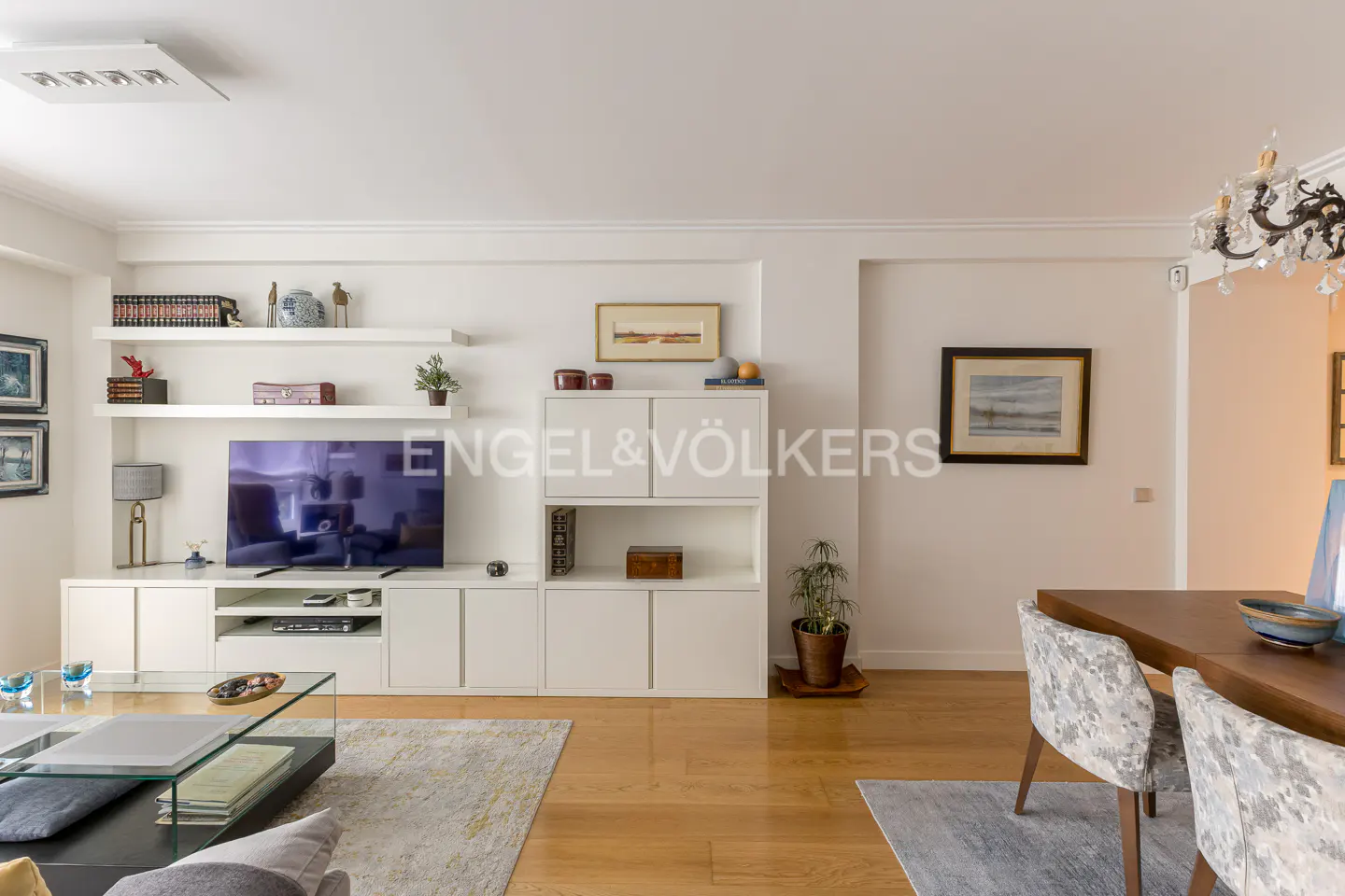 Modern living room with white media console, flat-screen TV, floating shelves, wooden floors, and a dining area.