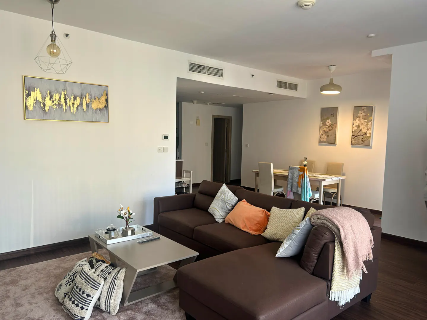 A living room with a brown sectional sofa, a gray coffee table, and a dining table with white chairs. Artwork adorns the walls.
