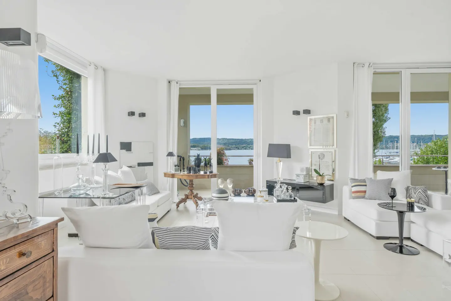 Bright, modern living room with white sofas, glass tables, and large windows overlooking a scenic harbor and blue sky.