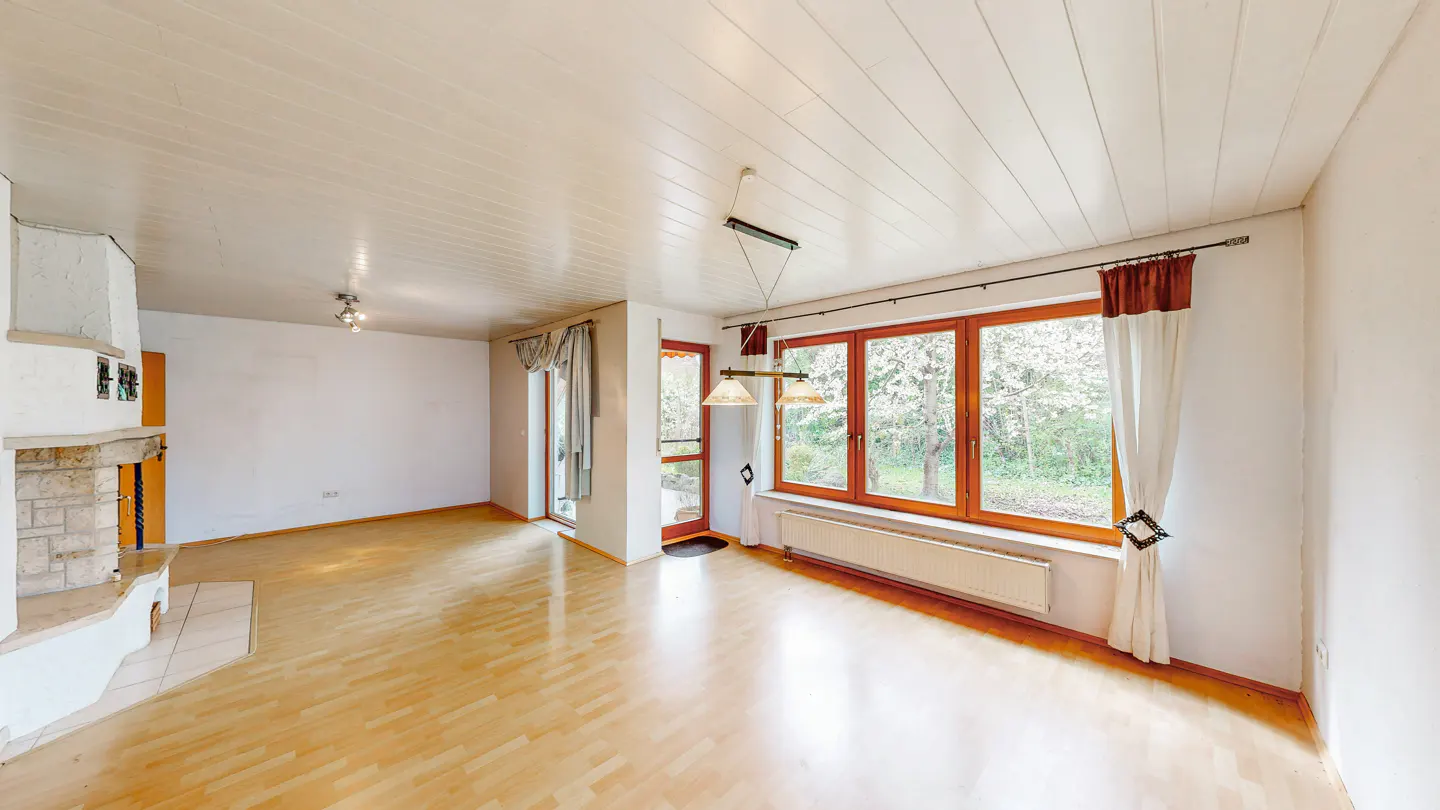 Bright living room with wood floors, white walls, and a wood-burning fireplace. Large windows offer a view of trees.