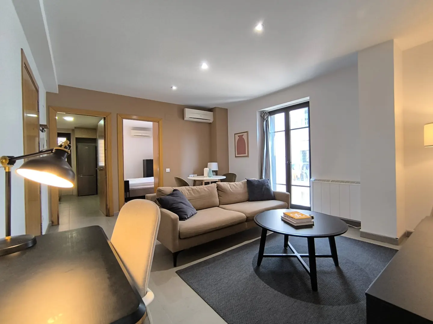 Modern living room with a beige sofa, black coffee table, desk with lamp, and views into a kitchen and bedroom.