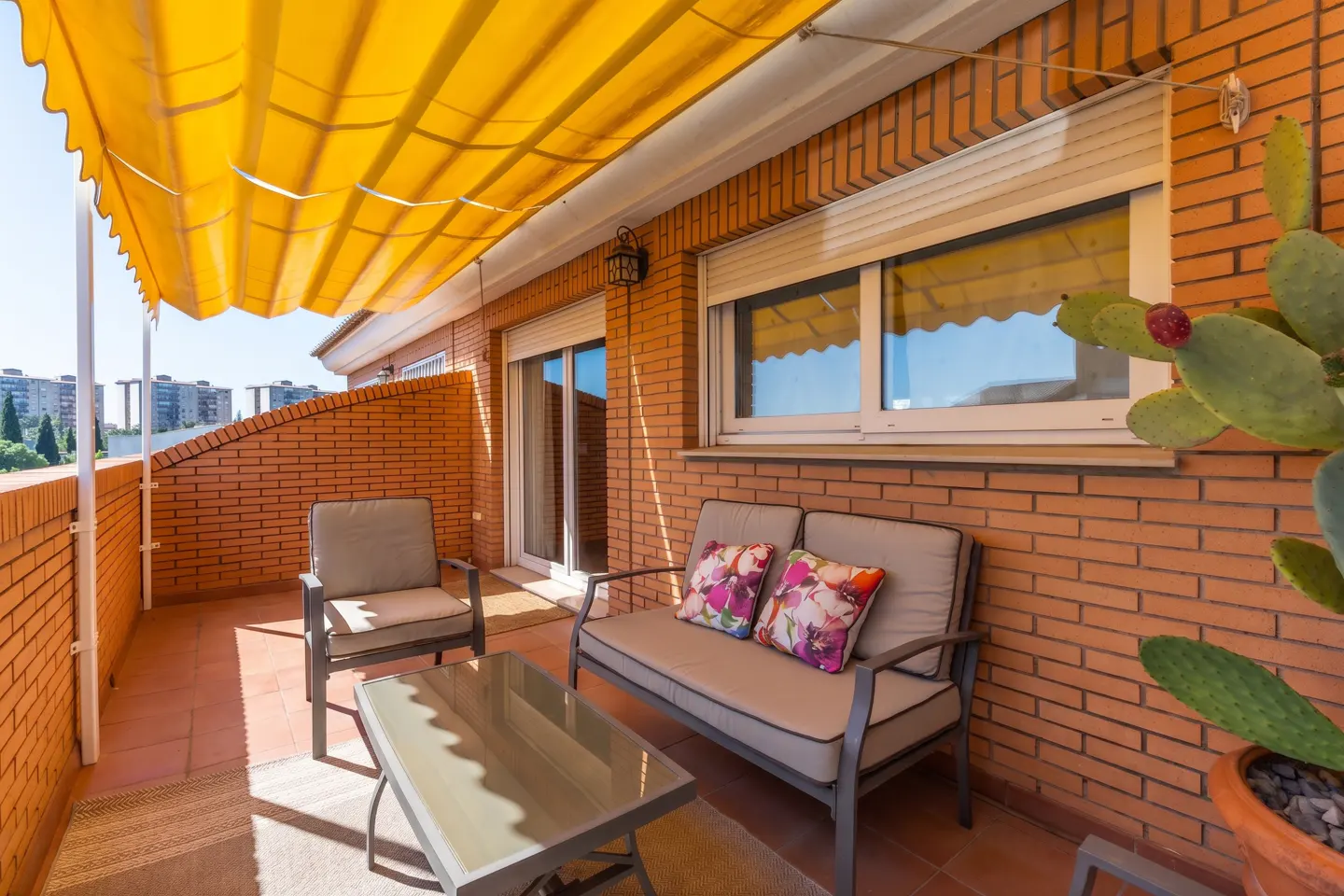 A brick balcony with a yellow awning features a loveseat, chair, and glass table. A cactus sits in a pot near a window.