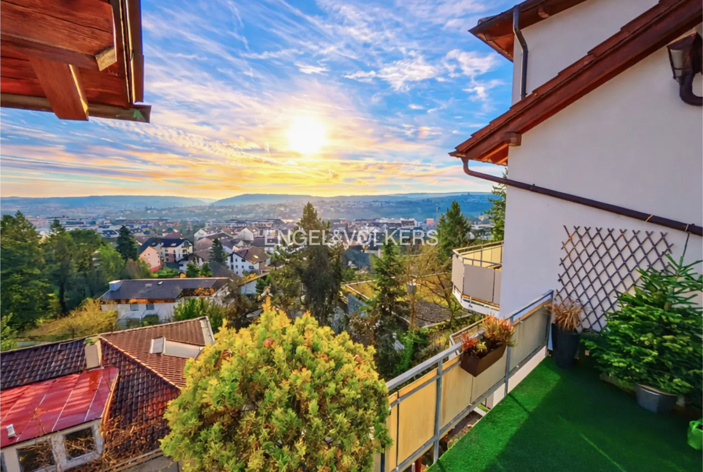 View from a balcony with green turf, overlooking a town at sunset. White building with brown roof on the right.