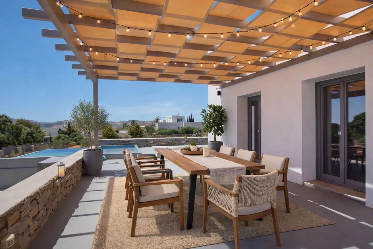 Outdoor dining area with a wooden table, chairs, and string lights under a pergola, overlooking a pool and landscape.