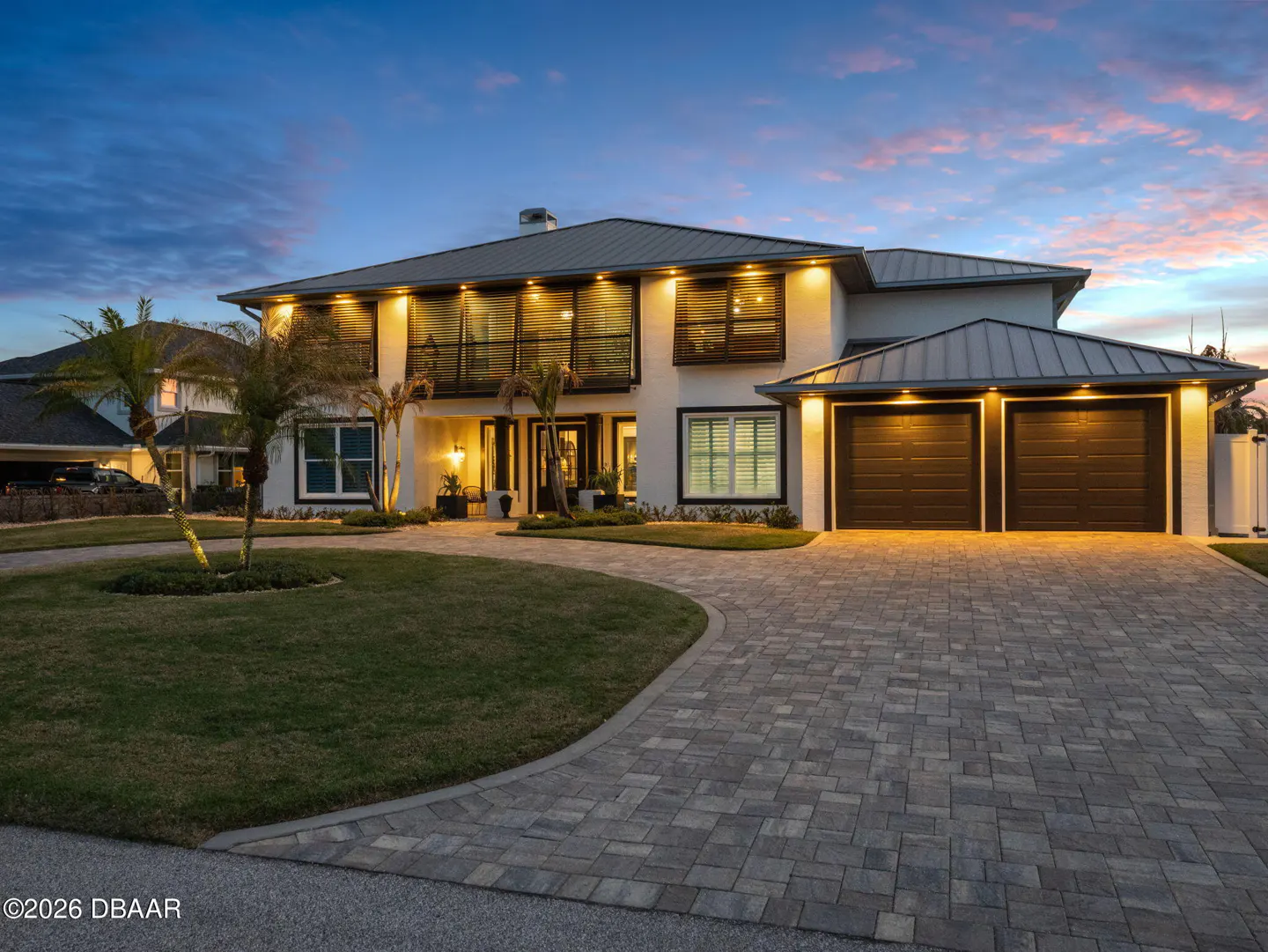 Two-story white house with a gray roof, black trim, and a two-car garage at dusk. A brick driveway leads to the house.
