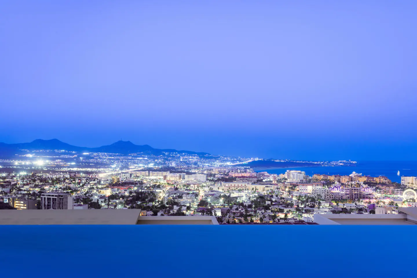 Night view of a city from a rooftop pool, with mountains and the ocean in the background. The sky is a deep blue.