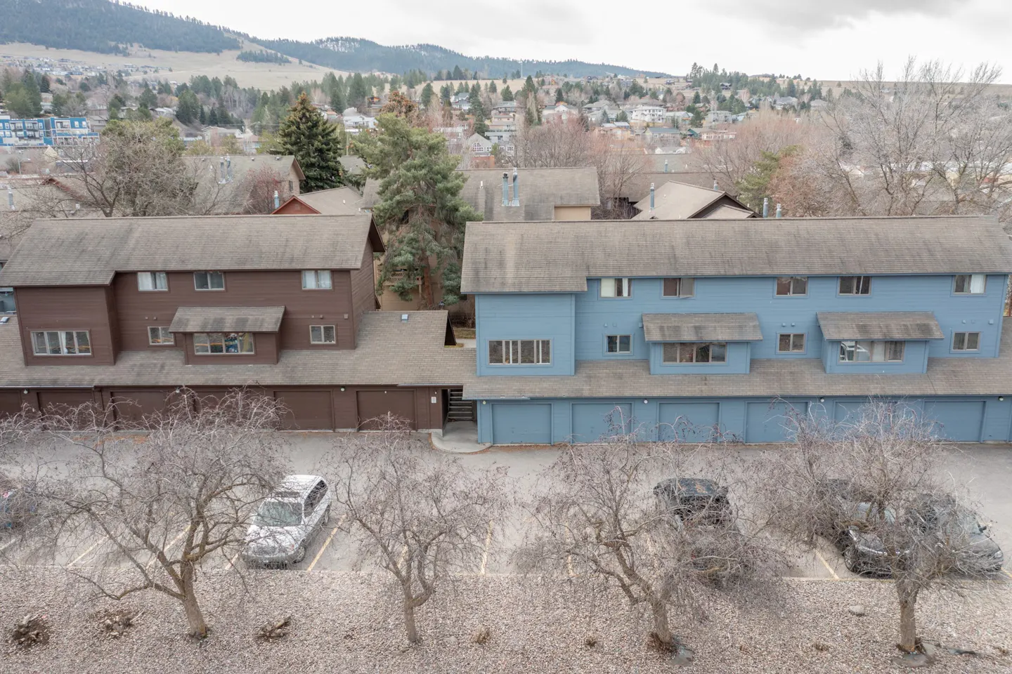 Aerial view of brown and blue two-story apartments with garages, parked cars, and bare trees in front of a hillside neighborhood.
